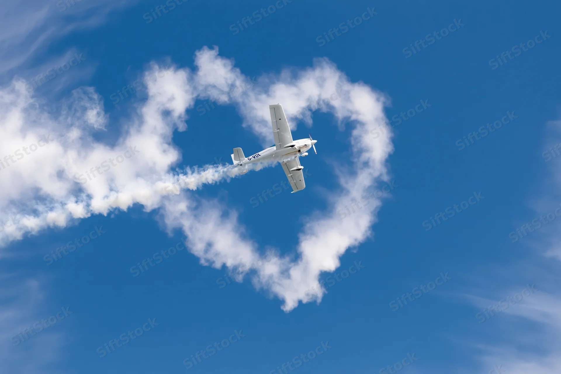 Small white propeller plane flying through clear blue sky leaving heart-shaped smoke trail