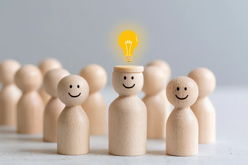 Wooden figurines in a group with one having a glowing lightbulb above its head symbolizing an idea