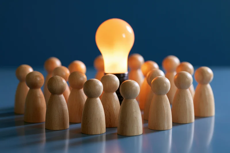 Wooden peg figures arranged in a circle around a glowing lightbulb on a reflective blue surface