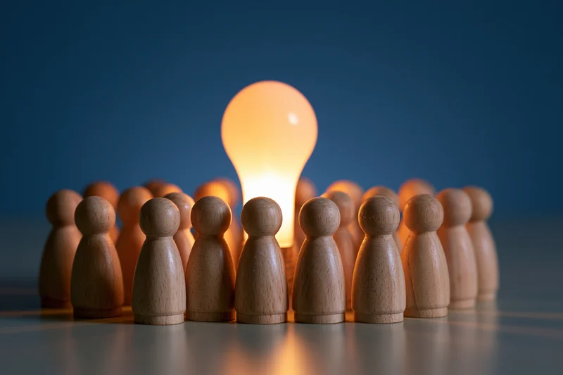Wooden figures arranged in a circle around a glowing lightbulb on a reflective surface