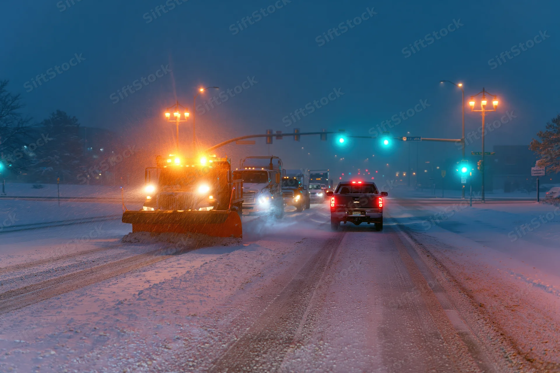 Snowplow truck clearing snow on a city street at night with traffic and streetlights
