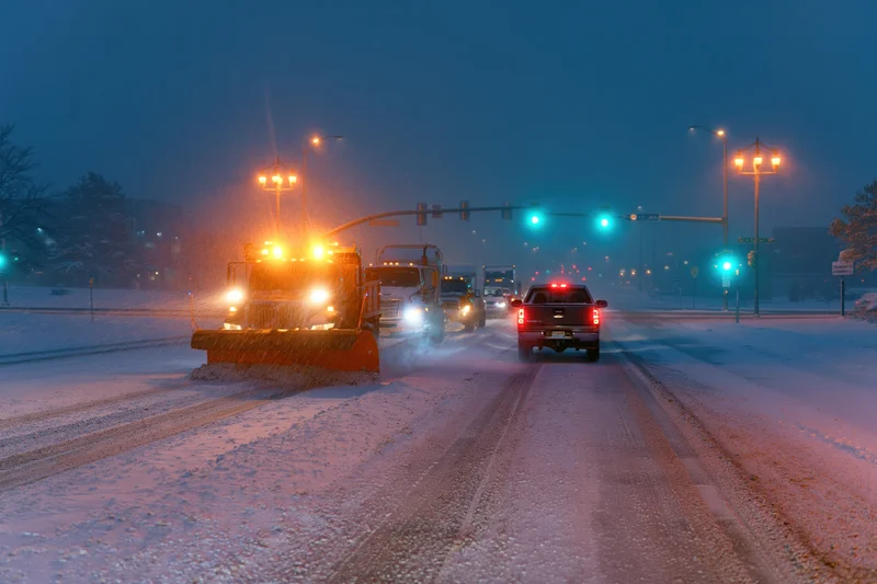 Snowplow truck clearing snow on a city street at night with traffic and streetlights