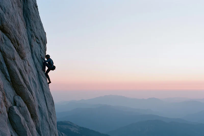 Solo climber ascending steep granite cliff at dusk with mountain range in background