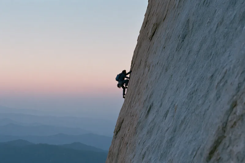 Solo climber ascending steep granite cliff at dusk with mountain valley in background