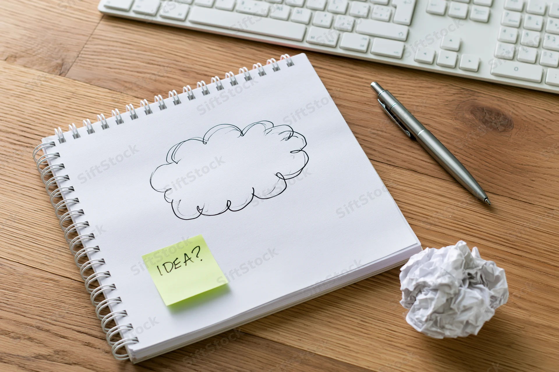 Spiral-bound notebook with hand-drawn cloud and sticky note labeled idea on wooden desk beside keyboard and pen