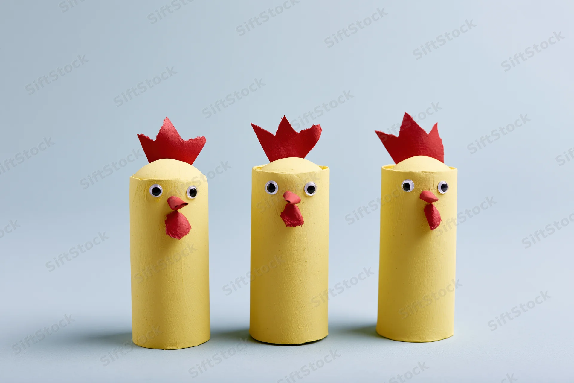 Three handmade yellow cardboard tube chickens with red combs and eyes arranged in a row on a plain light background