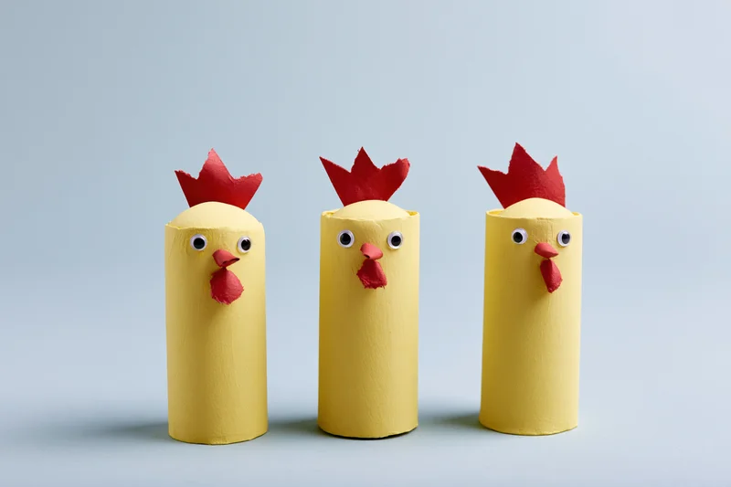 Three handmade yellow cardboard tube chickens with red combs and eyes arranged in a row on a plain light background