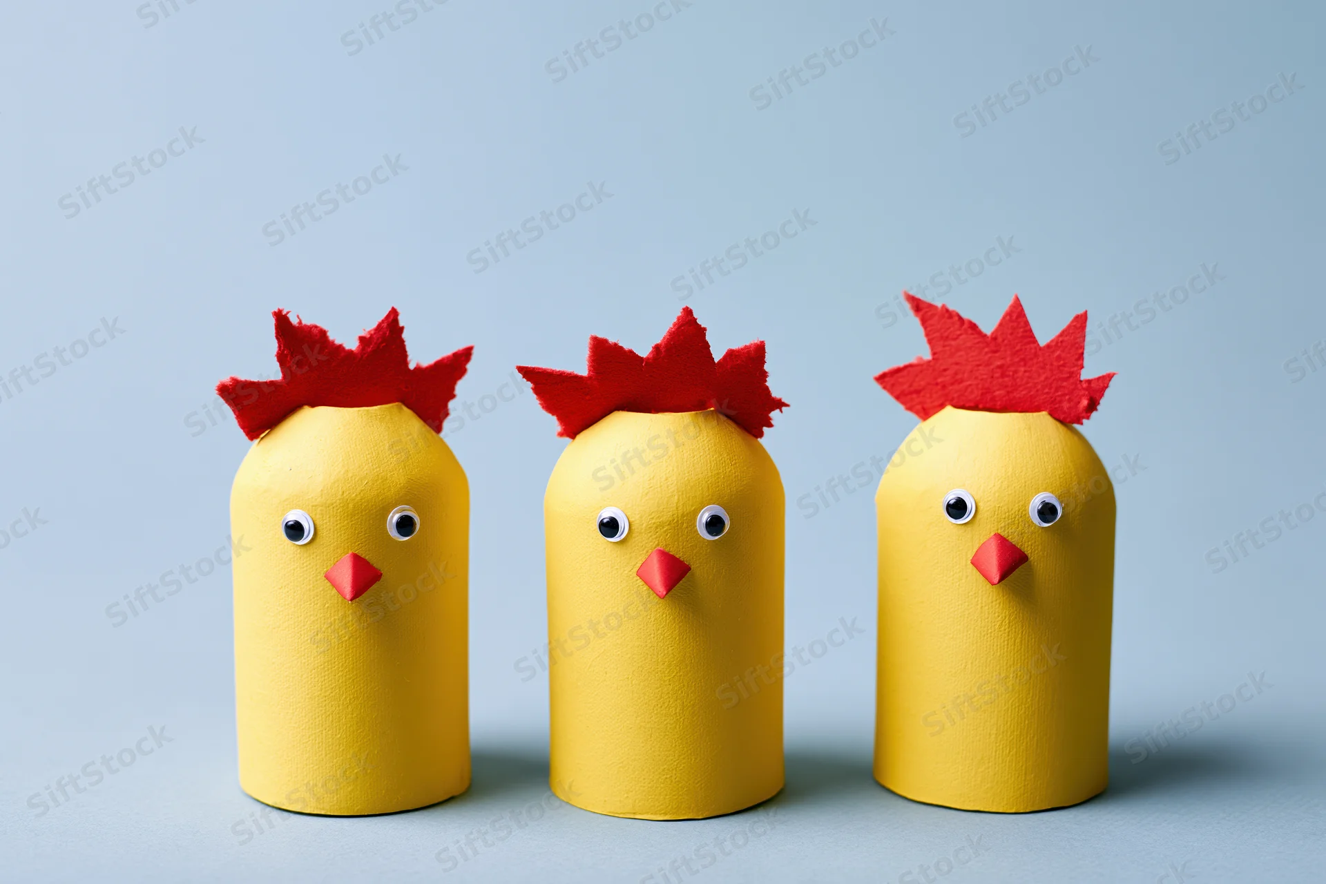 Three handmade yellow cardboard tube chicks with red felt combs and eyes arranged in a row on a blue surface