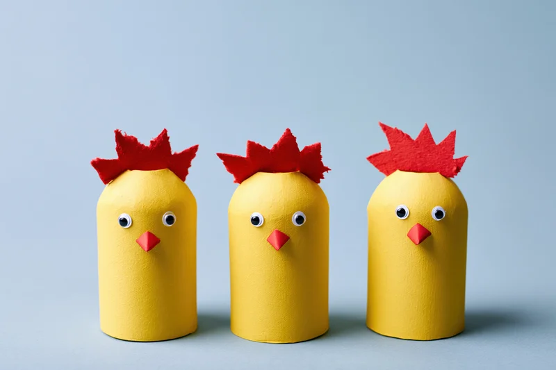 Three handmade yellow cardboard tube chicks with red felt combs and eyes arranged in a row on a blue surface