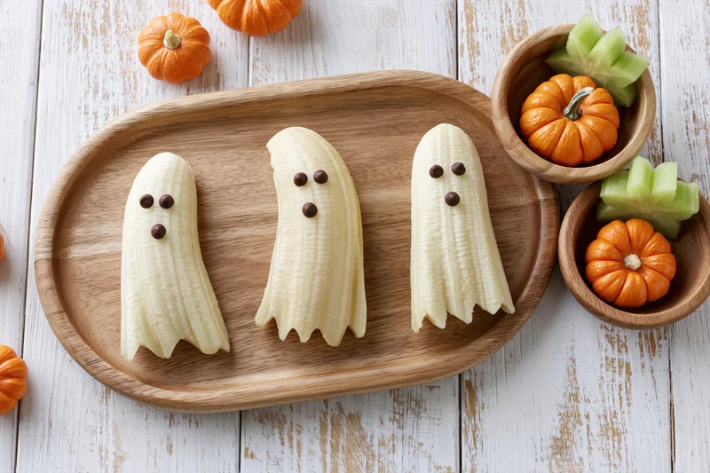 Three peeled bananas shaped like ghosts with chocolate chip eyes on a wooden tray surrounded by small pumpkins and melon slices on a white rustic table