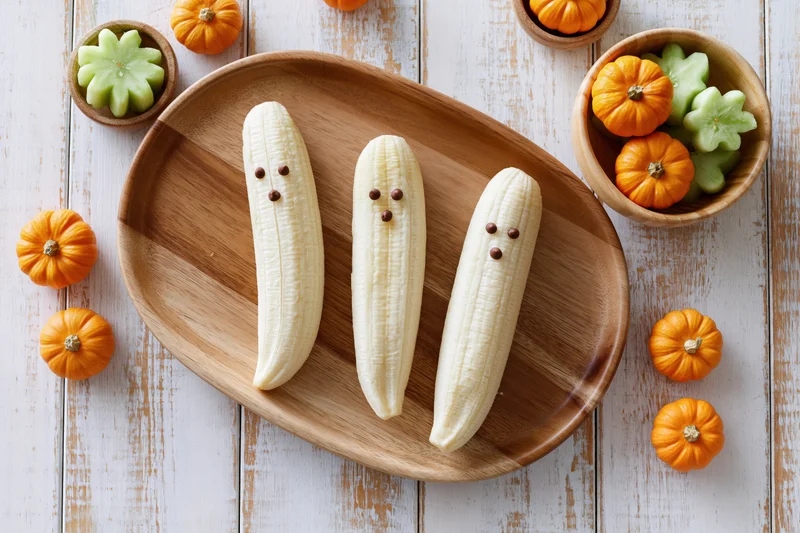 Three peeled bananas with chocolate chip faces on a wooden plate surrounded by small pumpkins and green candies on a white wooden table
