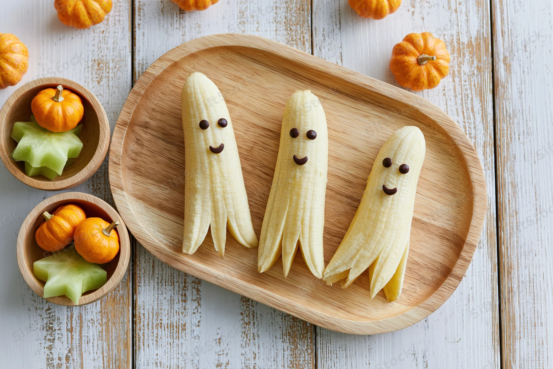 Three peeled bananas with chocolate chip faces arranged on a wooden tray with small pumpkins and celery stars nearby