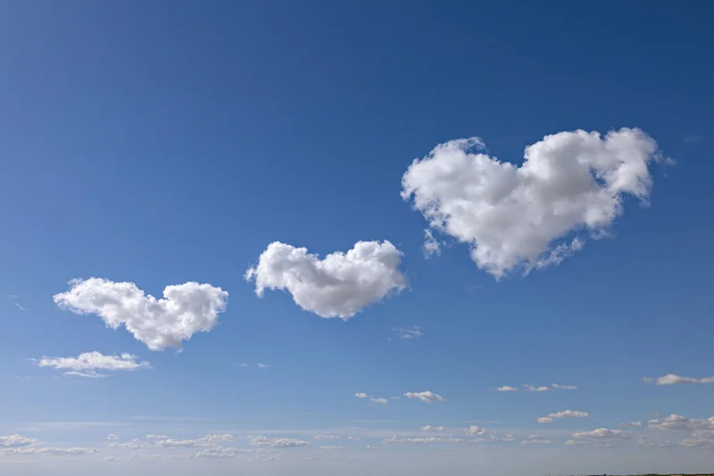 Three heart-shaped clouds floating in a clear blue sky over a distant horizon