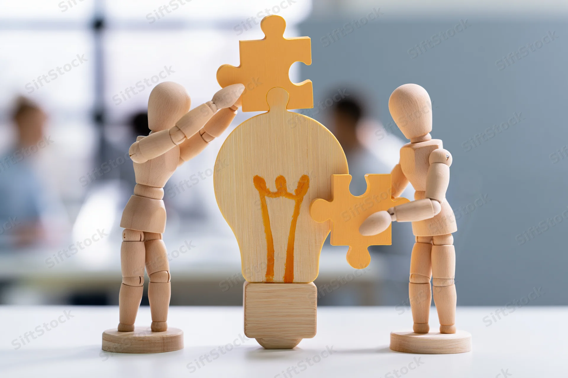 Wooden mannequins assembling puzzle pieces around a lightbulb on a desk in an office setting