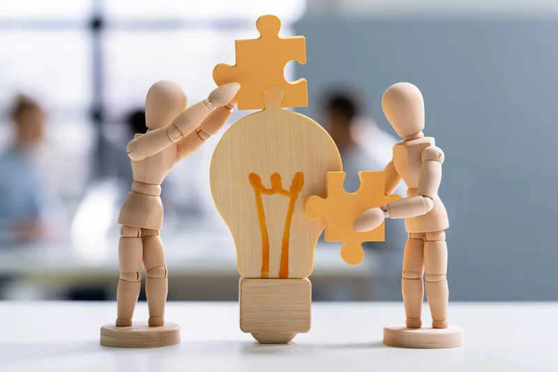 Wooden mannequins assembling puzzle pieces around a lightbulb on a desk in an office setting