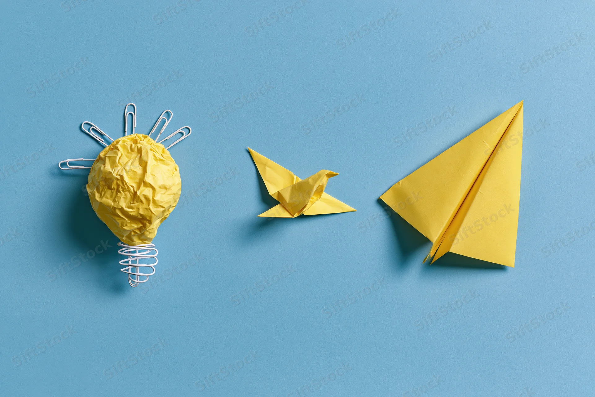 Three yellow paper origami objects — a lightbulb, bird, and airplane — arranged in a row on a blue background