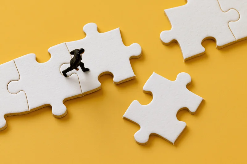 Miniature businessman figurine walking across white puzzle pieces on yellow background
