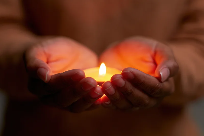 Cupped hands holding a small glowing candle in a dimly lit setting