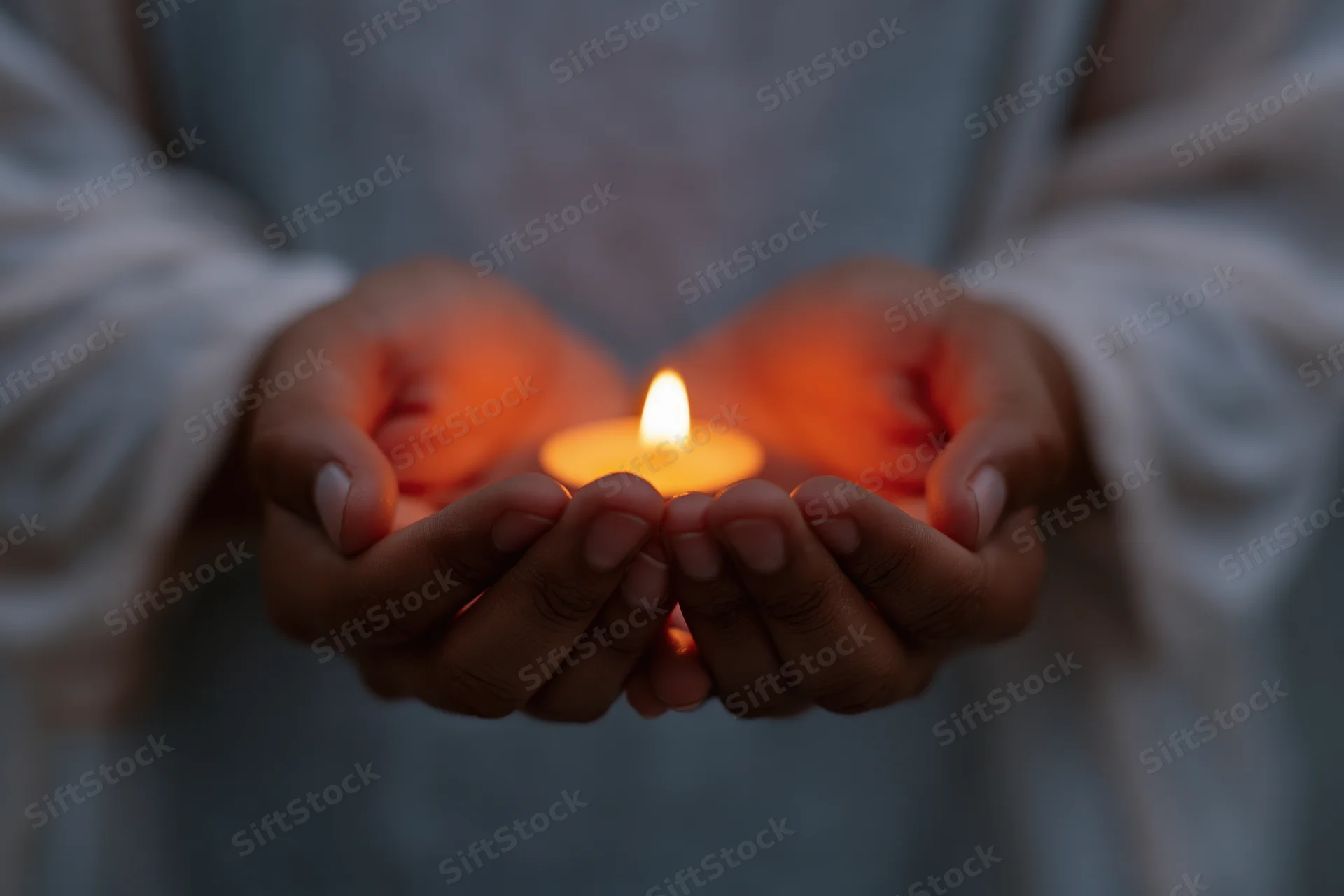Cupped hands holding a small lit candle in a soft glow