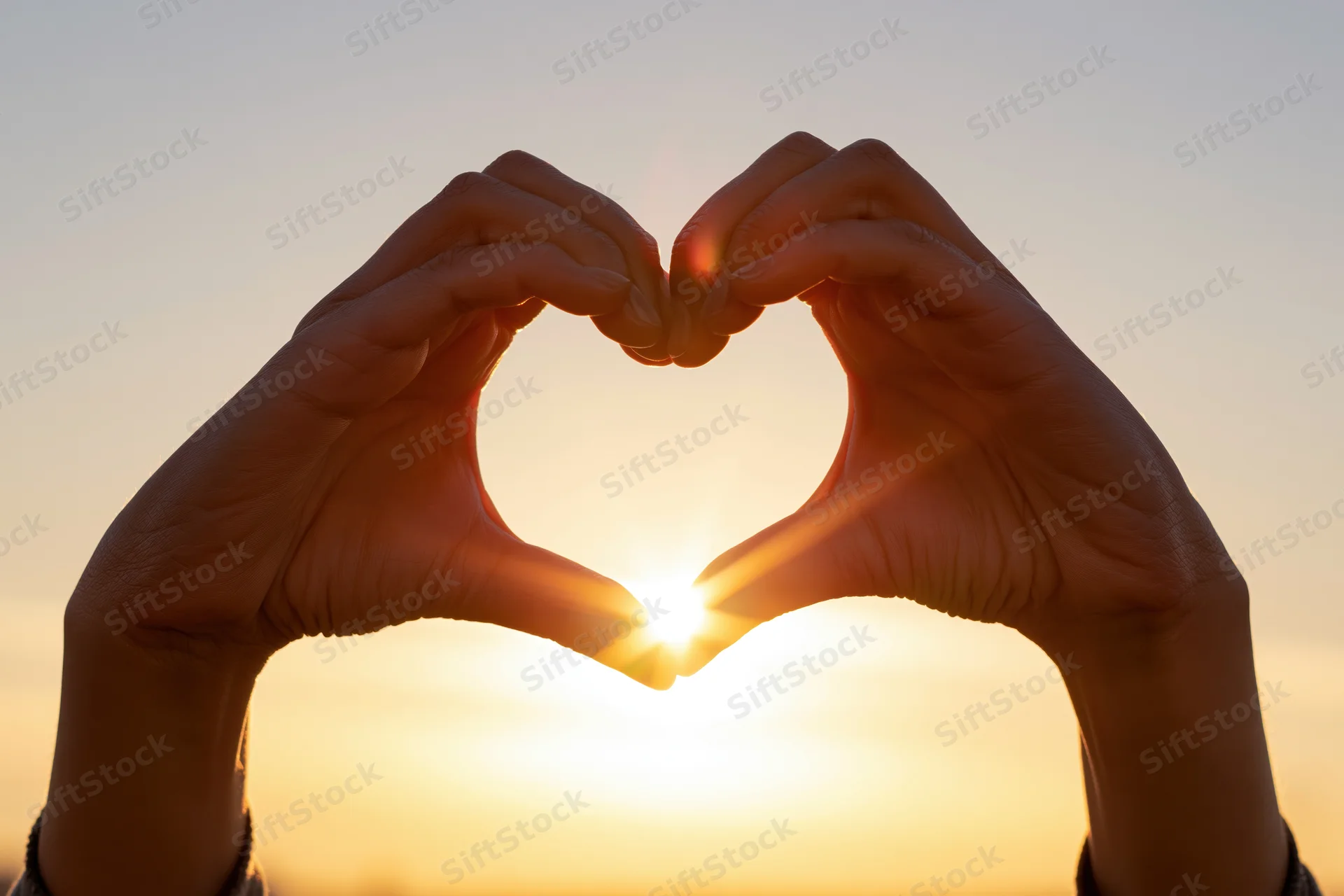 Hands forming heart shape against sunset sky