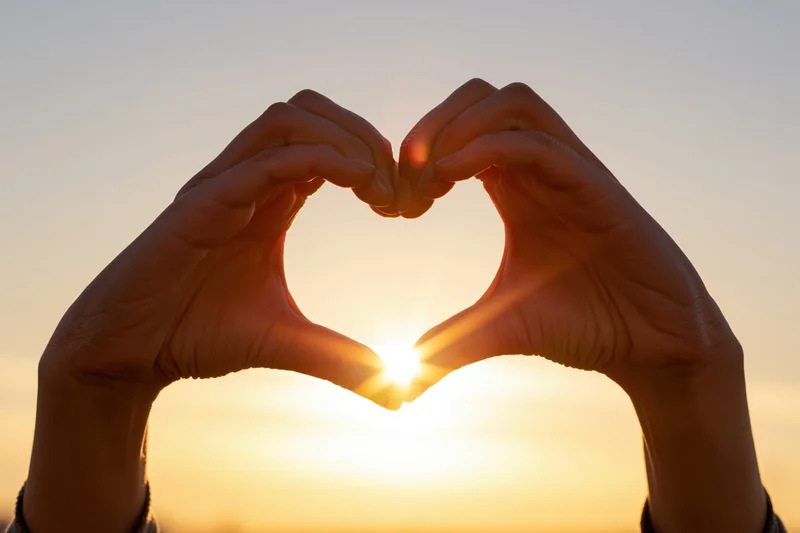 Hands forming heart shape against sunset sky