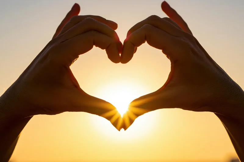 Hands forming heart shape against sunset sky