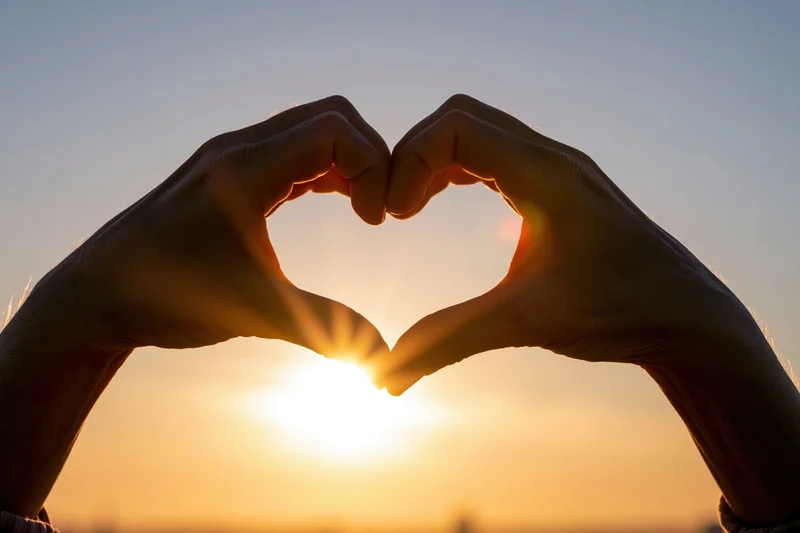 Hands forming heart shape against sunset sky