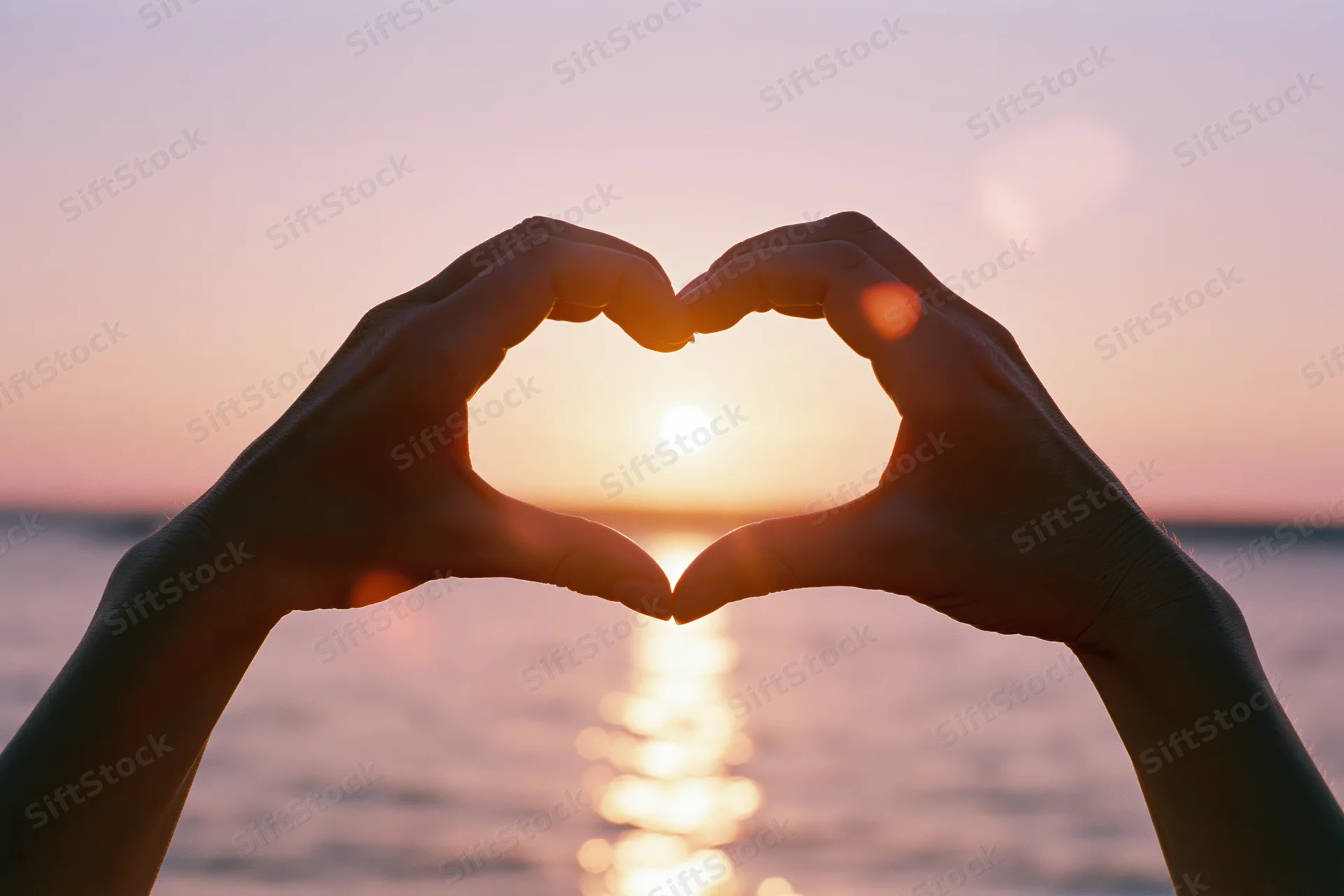 Hands forming a heart shape against a glowing sunset over the ocean