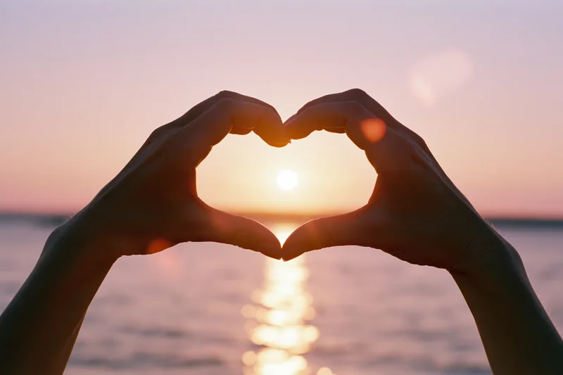 Hands forming a heart shape against a glowing sunset over the ocean