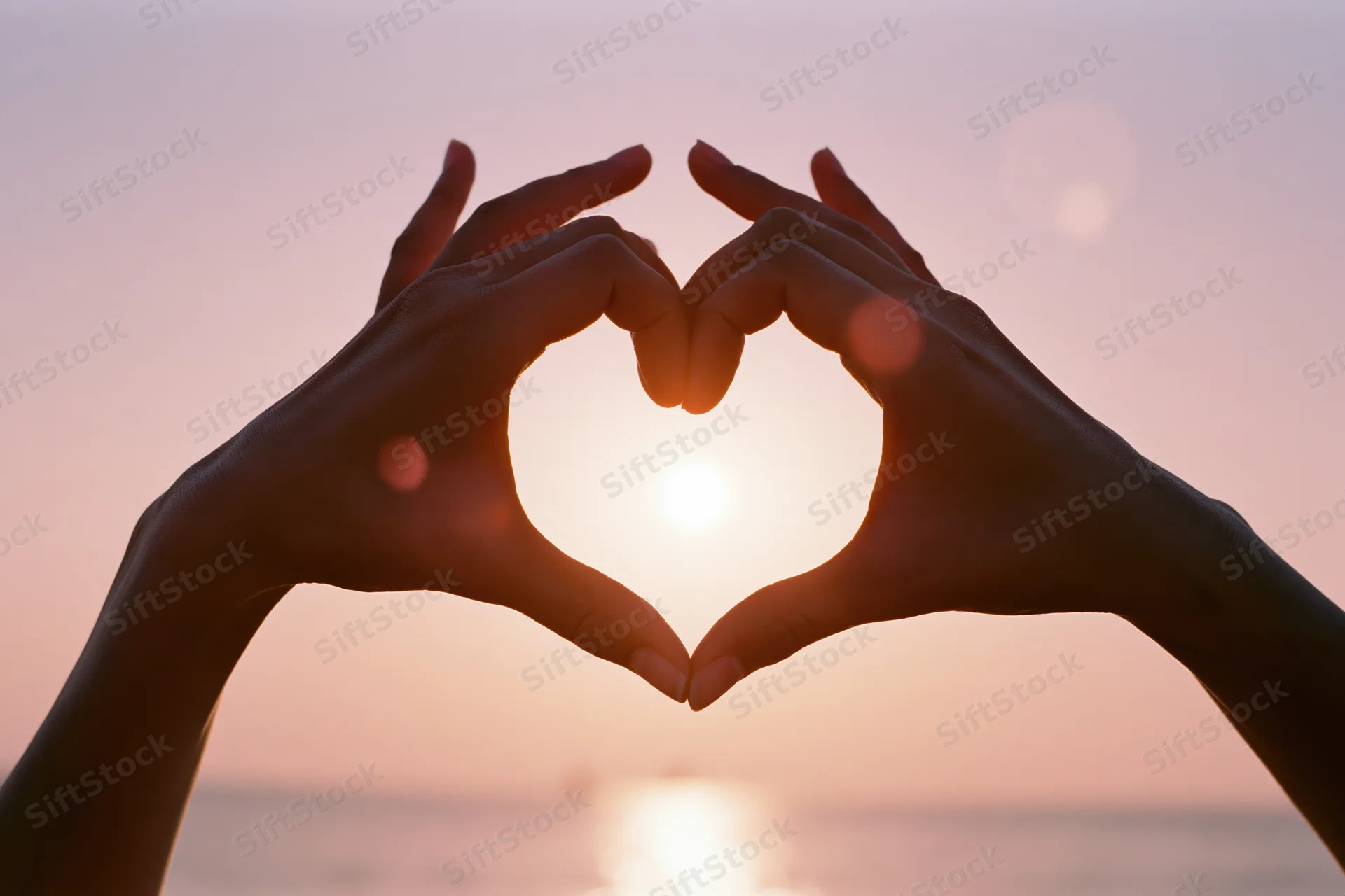 Hands forming a heart shape against a glowing sunset over the ocean