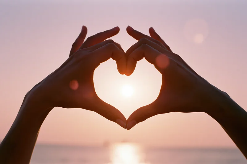 Hands forming a heart shape against a glowing sunset over the ocean