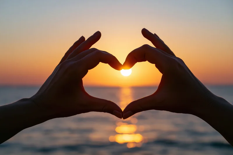 Hands forming heart shape silhouetted against sunset over ocean