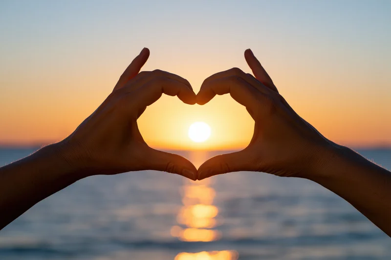 Hands forming heart shape silhouetted against sunset over ocean