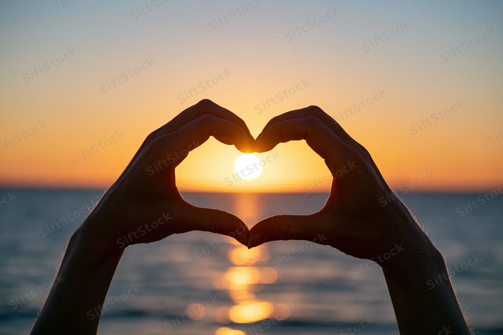 Hands forming heart shape silhouetted against sunset over ocean