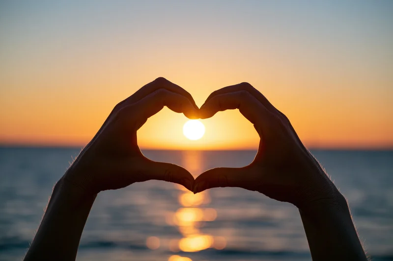 Hands forming heart shape silhouetted against sunset over ocean
