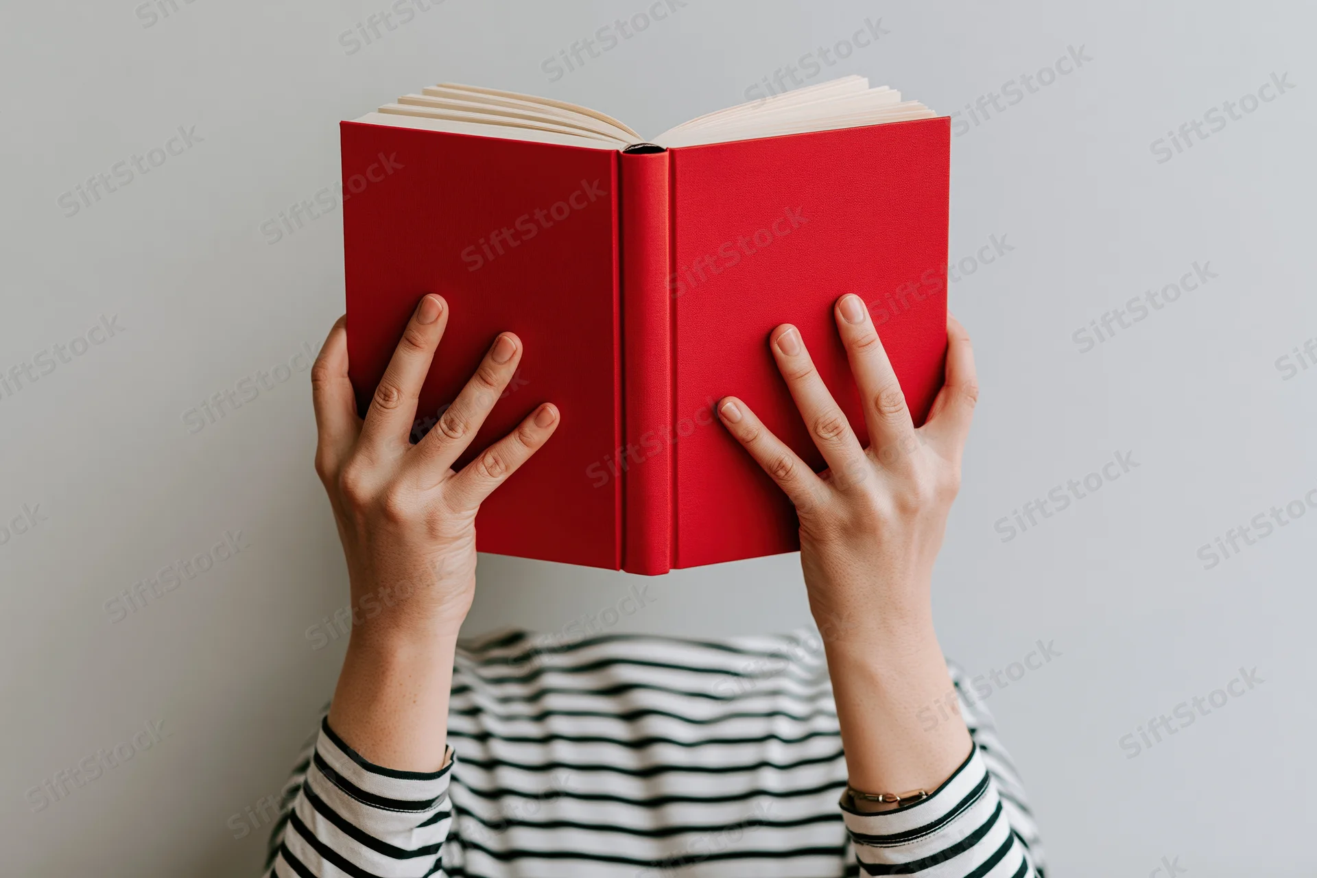 Woman holding open red hardcover book above her head, striped shirt visible below, plain gray background
