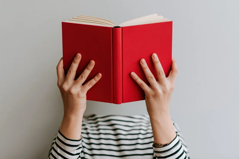 Woman holding open red hardcover book above her head, striped shirt visible below, plain gray background