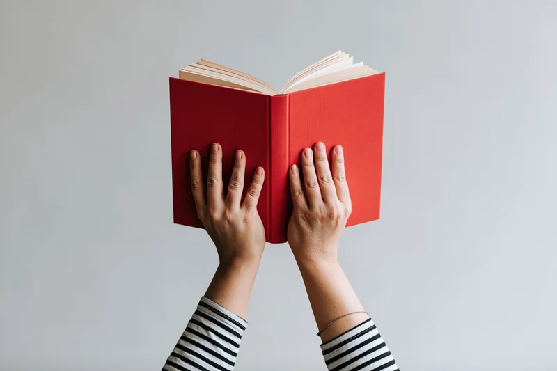Hands holding open red hardcover book above head against plain background