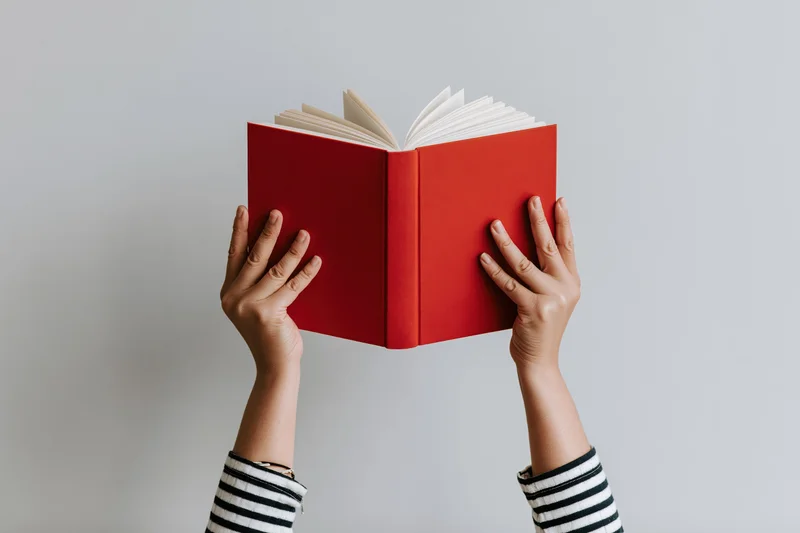 Hands holding open red hardcover book above head against white background