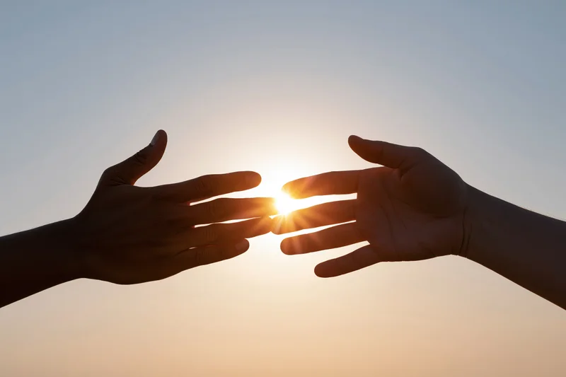 Two hands reaching toward each other against a bright backlit sky with the sun glowing between them