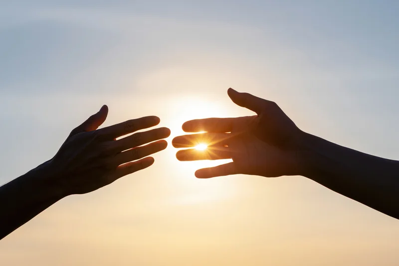 Two hands reaching toward each other against a bright backlit sky at sunrise