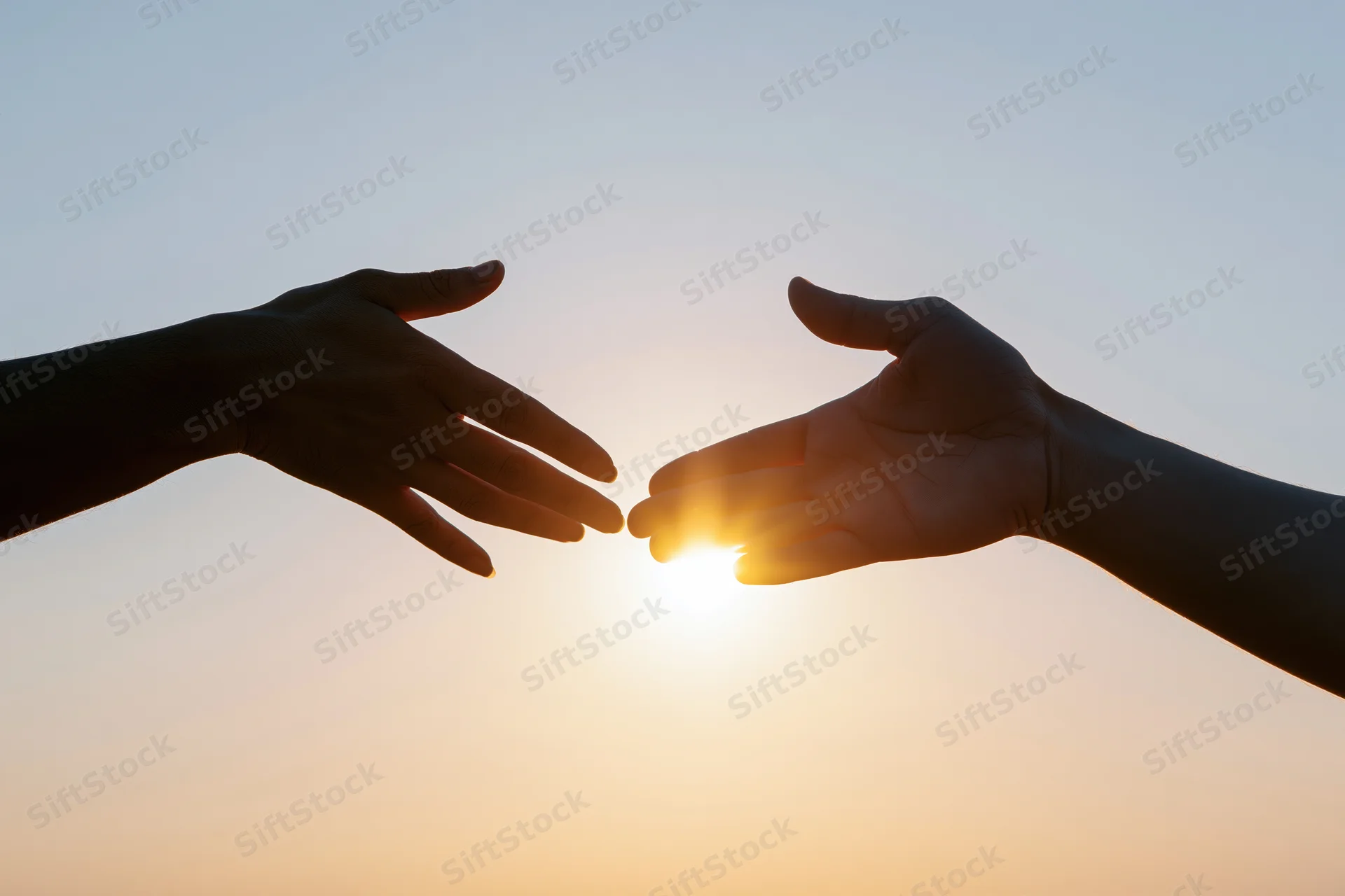 Two hands reaching toward each other against a bright backlit sky at sunset