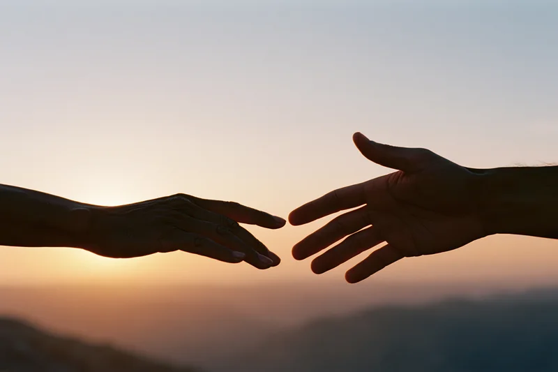 Two hands reaching toward each other in silhouette against a sunset sky