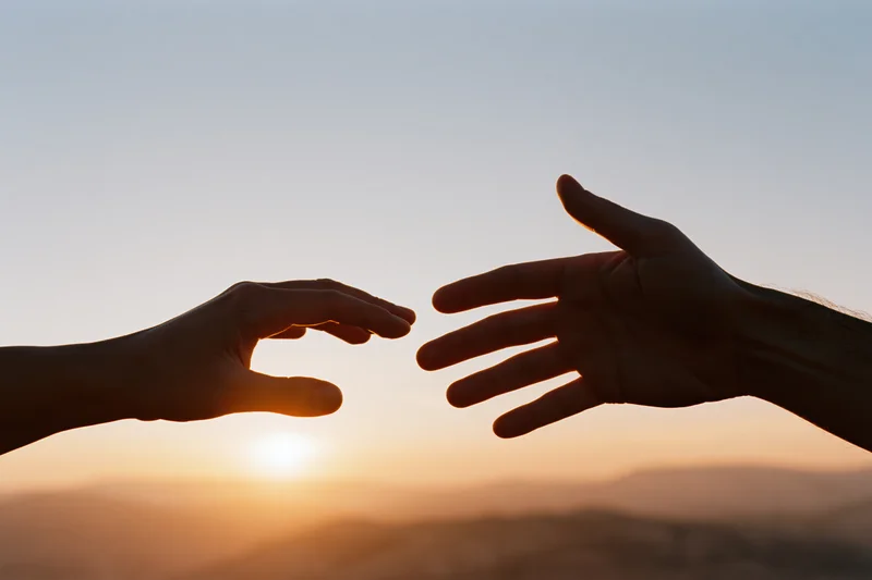 Two hands reaching toward each other in silhouette against a sunset sky