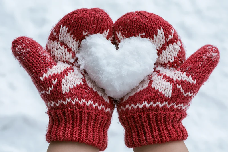 Hands in red knitted mittens holding a heart-shaped snowball in snowy winter setting