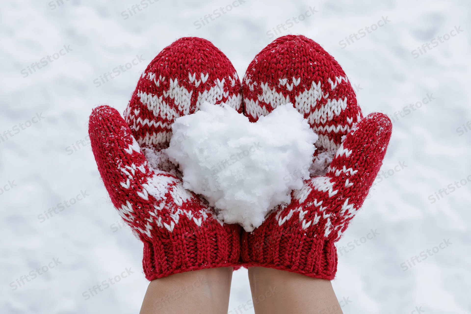 Hands in red knitted mittens holding a heart-shaped snowball in snowy winter setting