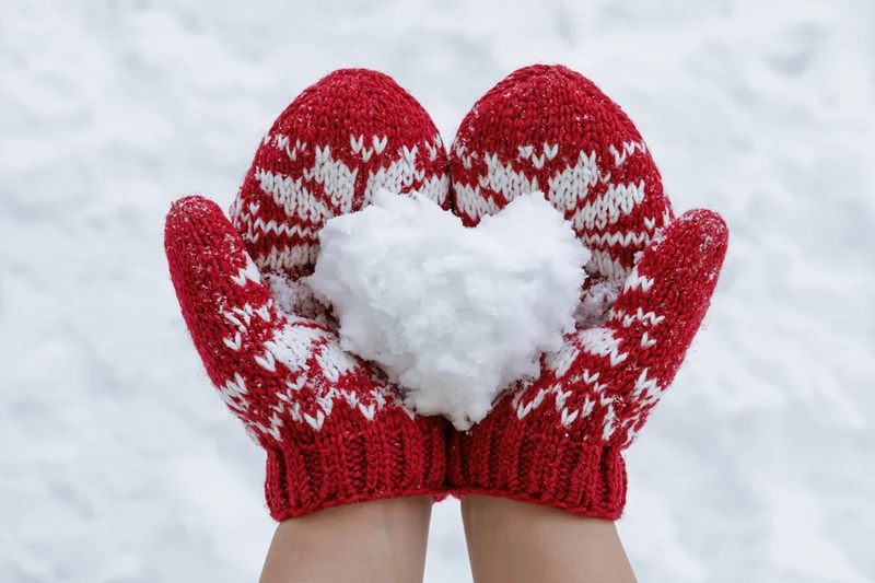 Hands in red knitted mittens holding a heart-shaped snowball in snowy winter setting