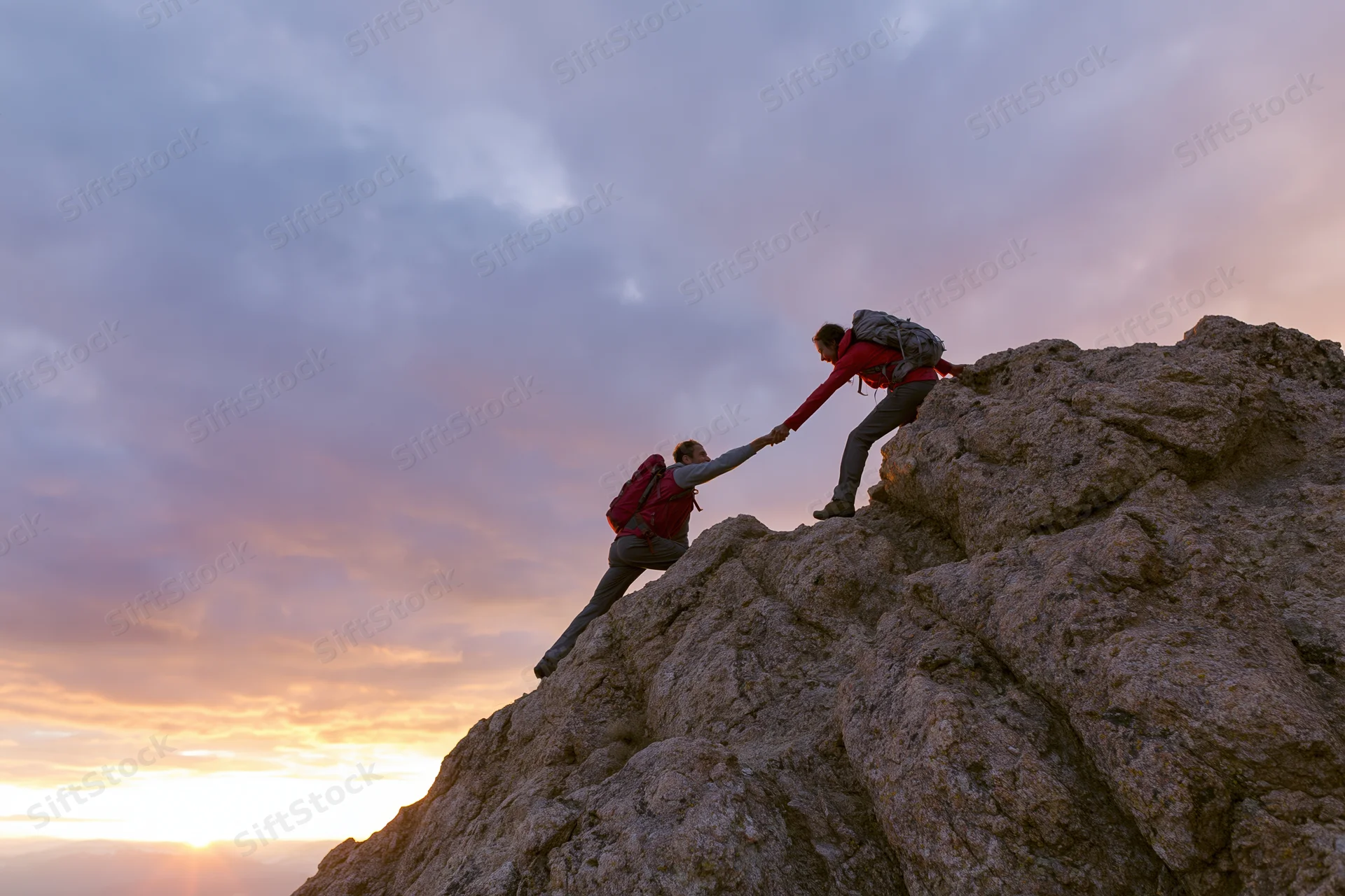 Two hikers at rocky summit with one pulling the other up by hand during sunset