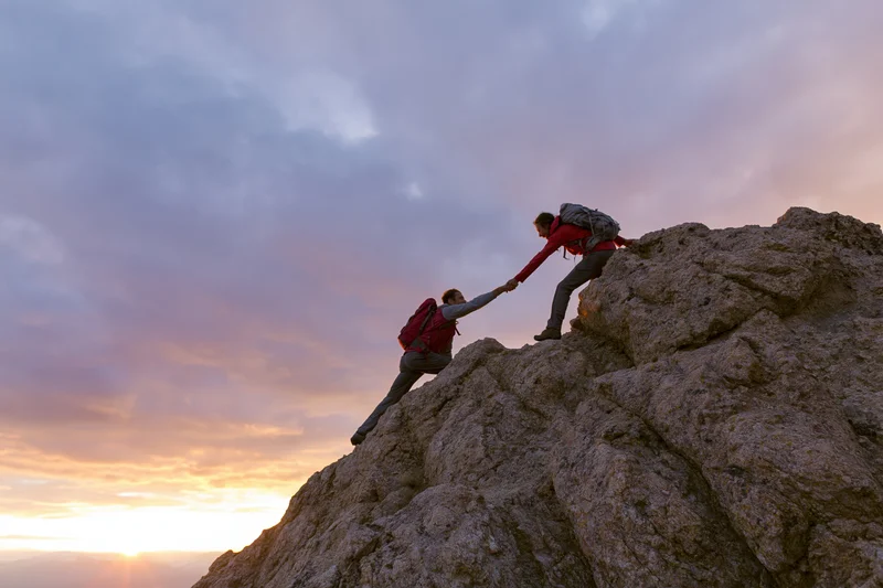 Two hikers at rocky summit with one pulling the other up by hand during sunset