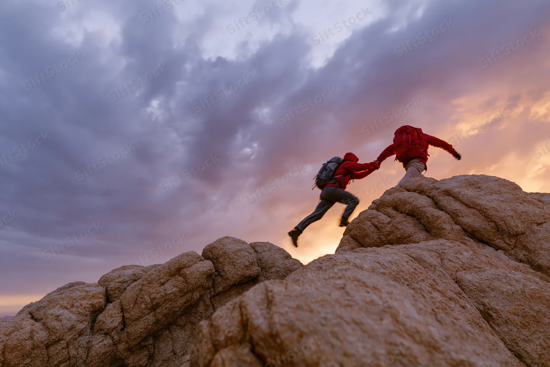 Two hikers helping each other climb rocky cliff at sunset
